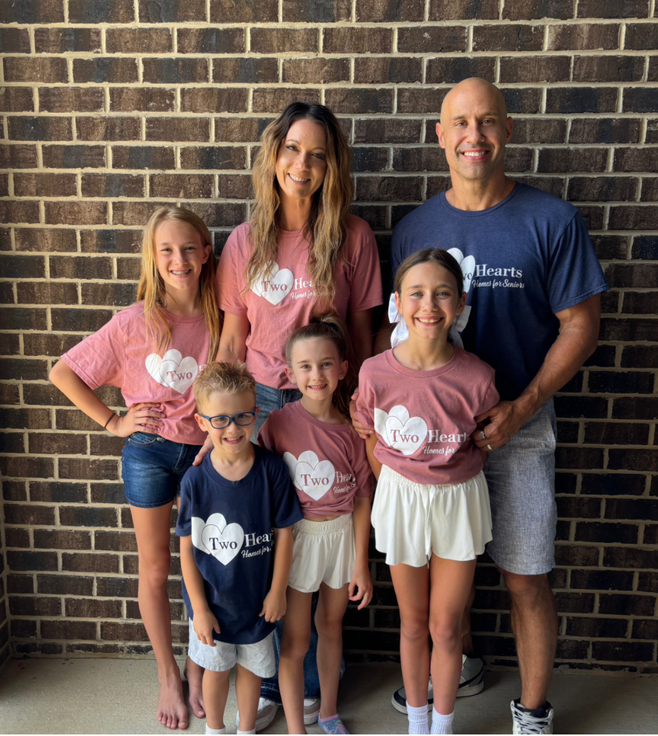 The family behind Two Hearts Homes for Seniors, smiling together in branded t-shirts, representing their commitment to compassionate, family-centered senior care.